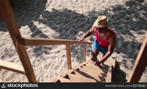 Handsome afro american man in sunglasses and in hat climbing up a wooden ladder on tree house at the beach and waving hello to his friends.