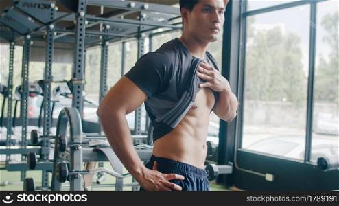 Handsome asian guy take out shirt naked torso body look at his muscles and trained body at mirror in fitness . Bodybuilder male model posing, Fitness in gym, sport and healthy lifestyle concept.