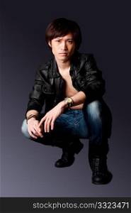 Handsome Asian male wearing leather jacket over a bare chest and jeans with macho attitude while crouching, isolated.