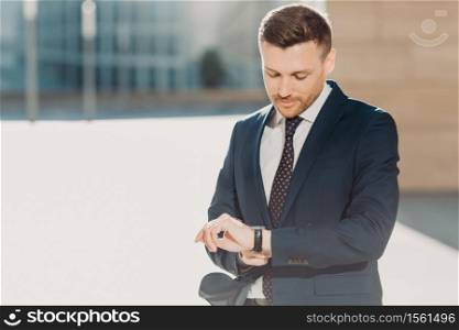 Handsome attractive male checks time, hurries for business meeting, looks at watch, dressed in formal black suit, poses outdoor near office building. People, business and occupation concept.