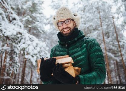 Handsome bearded male in eyewear and warm hat with anorak, holds firewood, poses against trees covered with white sparkling snow, spends time in forest. Outdoor shot of happy man rests in forest