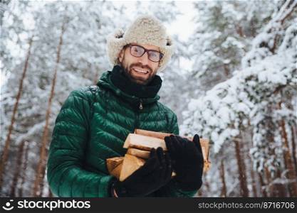 Handsome bearded male in eyewear and warm hat with anorak, holds firewood, poses against trees covered with white sparkling snow, spends time in forest. Outdoor shot of happy man rests in forest