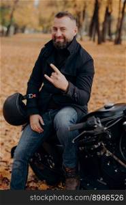 Handsome bearded male motorcyclist holds helmet, makes horn gesture with fingers, feels cool, wears black coat and jeans, sits on fast motorbike against autumn park background. Carefree racer