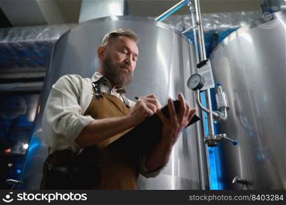Handsome bearded man brewer inside modern beer factory around steel tanks. Mature male worker making notes about technological process. Handsome bearded man brewer inside modern beer factory around steel tanks