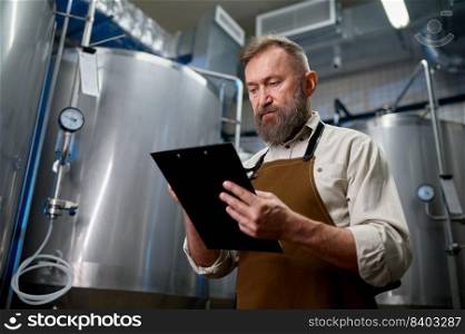 Handsome bearded man brewer inside modern beer factory around steel tanks. Mature male worker making notes about technological process. Handsome bearded man brewer inside modern beer factory around steel tanks