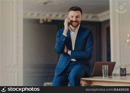 Handsome bearded man in blue suit, satisfied with good news, talking on the phone, posing at desk with modern laptop. Mobile conversation.