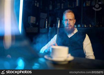 Handsome bearded man sitting at table and smoking hookah while drinking coffee in bar. Selective focus. Handsome bearded man smoking hookah while drinking coffee in bar