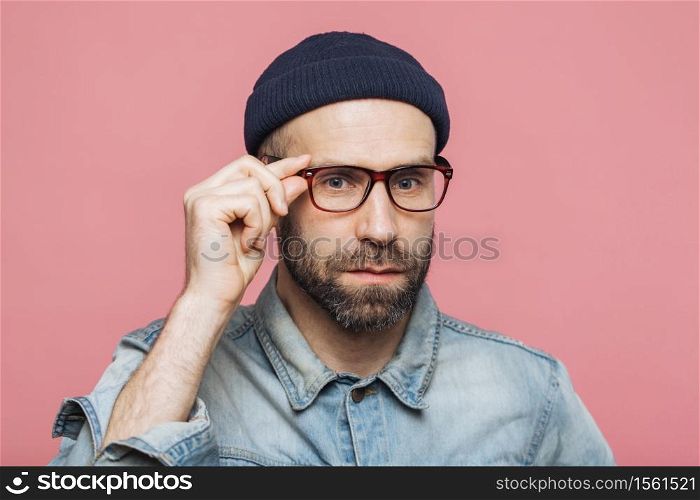 Handsome bearded man with serious expression wears spectacles and glasses, dressed in denim fashionable shirt, isolated over pink background. People, facial expressions and emotions concept.