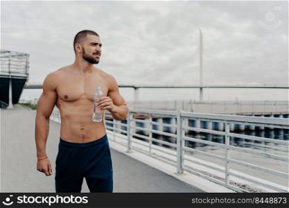 Handsome bearded sportsman drinks water from bottle promots healthy lifestyle and concentrated away into distance, relaxes after morning run, poses outside near river, being thirsty and tired.