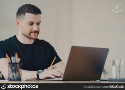 Handsome bearded young freelancer or office worker in casual wear holding pencil watching webinar or surfing internet sitting at table while working on modern laptop remotely from home. Handsome young freelancer working on laptop remotely from home
