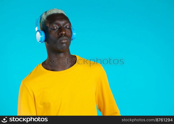 Handsome black man with trendy hairdo and headphones in studio against blue background. Guy in yellow outfit. Music, dance, radio concept. Handsome black man with trendy hairdo and headphones in studio against blue background. Guy in yellow outfit. Music, dance, radio concept.