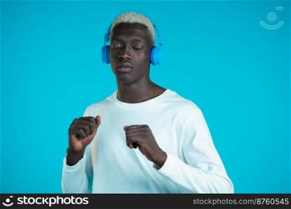 Handsome black man with trendy hairdo having fun, smiling, dancing with headphones in studio against blue background. Music, dance, radio concept. Handsome black man with trendy hairdo having fun, smiling, dancing with headphones in studio against blue background. Music, dance, radio concept.