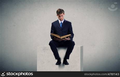 Handsome businessman sitting on white cube and reading book