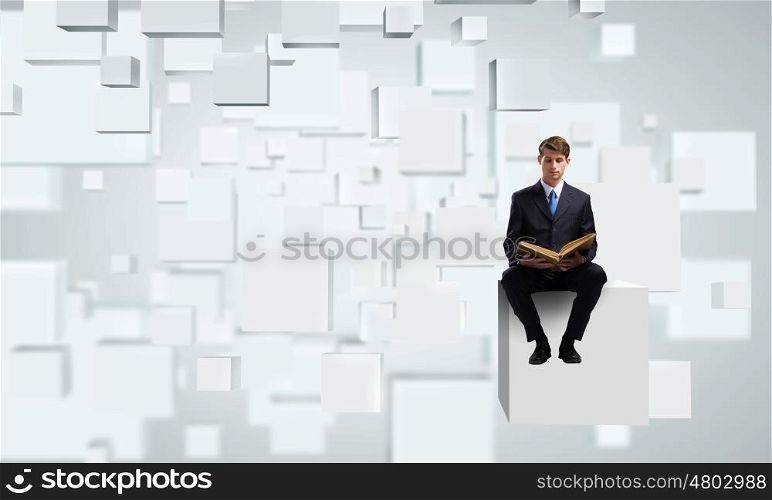 Handsome businessman sitting on white cube and reading book
