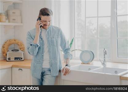 Handsome busy bearded businessman in casual comfy clothes standing in kitchen holding mobile phone, talking with client and dealing with customer issues while working remotely from home. Handsome busy bearded businessman in casual comfy clothes standing in kitchen with mobile phone