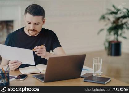 Handsome busy male freelancer or businessman in black tshirt analyzing document with information or sales report while working remotely from home, sitting at desk with laptop and other office suuplies. Focused entrepreneur analyzing document, working on project report in modern office