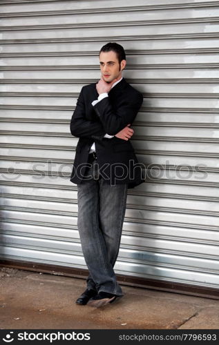 Handsome casual business man standing in front of and leaning against silver metal garage door with hand on chin thinking