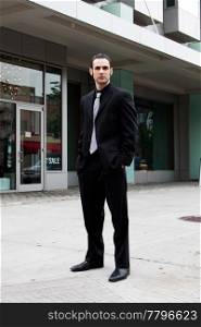Handsome Caucasian business man in black suit and hands in pocket standing on street in front of a modern building