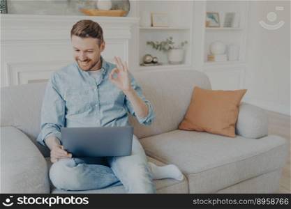 Handsome caucasian male freelancer smiling and showing sign OK with fingers while having video conference on notebook, young man in casual clothes using wireless laptop for online meeting from home. Handsome caucasian male freelancer smiling while talking online with boss on laptop