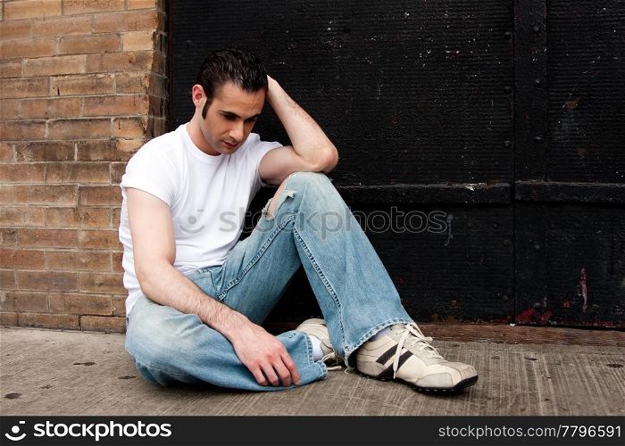 Handsome Caucasian man dressed in white shirt and blue jeans sitting on concrete floor in front of black metal door with a depressed expression thinking