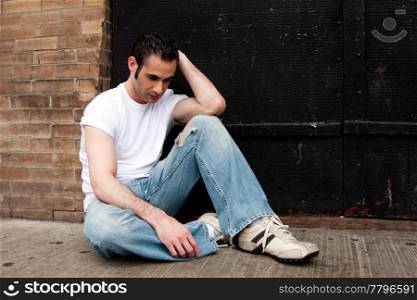 Handsome Caucasian man dressed in white shirt and blue jeans sitting on concrete floor in front of black metal door with a depressed expression thinking