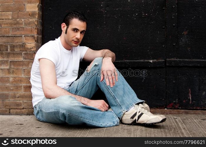 Handsome Caucasian man with sideburns dressed in white shirt and blue jeans sitting on concrete floor in front of black metal garage door