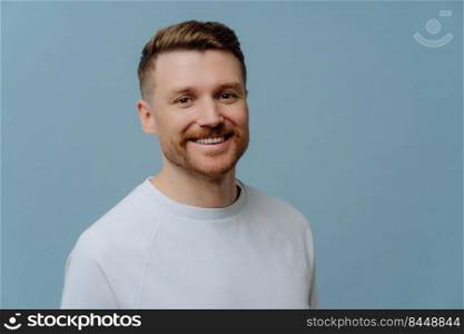 Handsome cheerful bearded man smiles gladfully dressed in casual white t shirt being in good mood poses against blue background with copy space. Glad European guy with stubble stands indoor.
