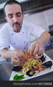 Handsome chef dressed in white uniform decorating pasta salad and seafood fish in modern kitchen