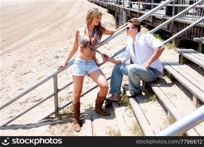 Handsome cool cute Caucasian guy flirting with sexy blond girl while sitting on some step that connect the boardwalk with the beach