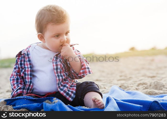 Handsome curious child sitting on sand on the beach playing, sunny day.