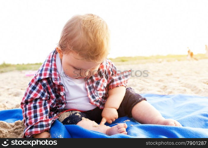 Handsome curious child sitting on sand on the beach playing, sunny day.