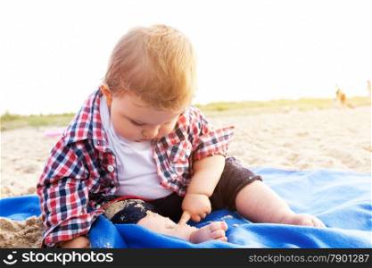 Handsome curious child sitting on sand on the beach playing, sunny day.