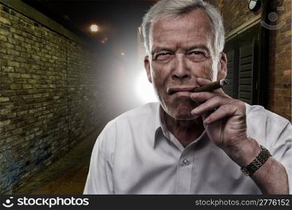 Handsome determined senior man smoking a cigar outdoors in the darkness backlit by a light on a brick building