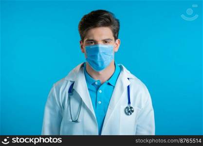 Handsome doctor man in professional white coat and medical mask on blue studio background. Serious doc looking to camera. High quality photo. Handsome doctor man in professional white coat and medical mask on blue studio background. Serious doc looking to camera