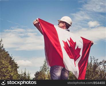 Handsome engineer, holding Canadian Flag in the park against the backdrop of green trees and the setting sun, looking into the distance. Close-up. Concept of labor and employment. Engineer, holding Canadian Flag in the park