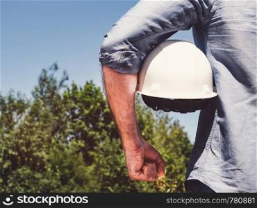 Handsome engineer, holding white hardhat in the park against the backdrop of green trees and the setting sun, looking into the distance. Close-up. Concept of labor and employment. Engineer, holding white hardhat in the park
