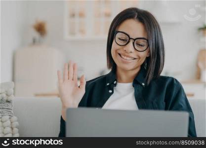 Handsome european girl in glasses has video call on computer and waving hand. Entrepreneur says hello to business partner. Distance work through internet on quarantine. Comfortable workplace at home.. Entrepreneur girl in glasses has video call on computer and waving hand. Distance work at home.