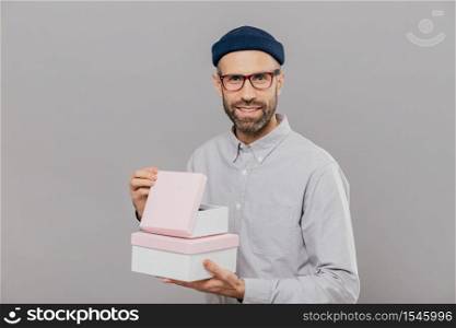 Handsome European man with thick stubble, pleasant smile, holds boxes in hands, opens gift with happiness, wears formal white shirt and headgear, isolated over grey background. Parcel in mans hands