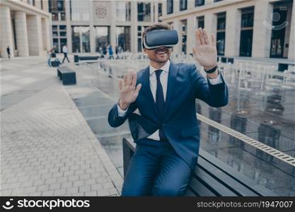 Handsome excited businessman sitting outside on bench while wearing portable Virtual Reality glasses, holding both of his hands in front of him, office buildings in blurred background. Handsome businessman sitting outside on bench while wearing portable Virtual Reality glasses