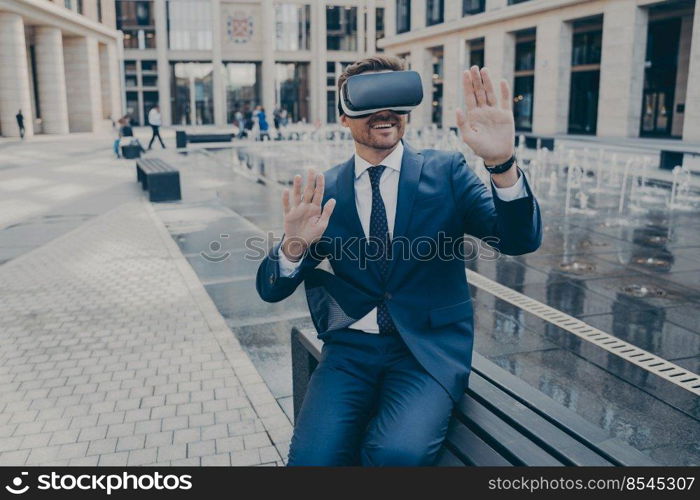 Handsome excited businessman sitting outside on bench while wearing portable Virtual Reality glasses, holding both of his hands in front of him, office buildings in blurred background. Handsome businessman sitting outside on bench while wearing portable Virtual Reality glasses