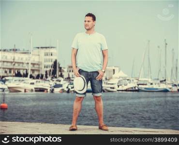 Handsome fashion man tourist on pier in port with yachts. Guy enjoying summer travel vacation by sea. Full length.