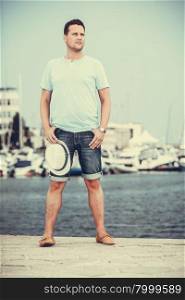 Handsome fashion man tourist on pier in port with yachts. Guy enjoying summer travel vacation by sea. Full length.