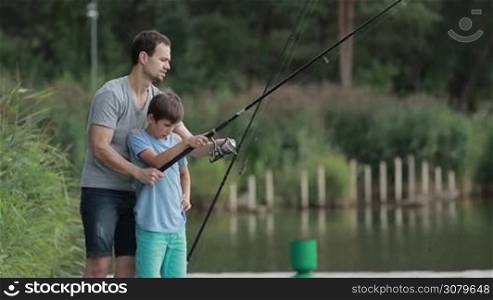 Handsome father teaching his teenage boy how to fish with fishing rod and reel on calm pond on sunny summer day. Side view. Affectionate dad and son relaxing in nature while fishing together at lake. Slow motion.
