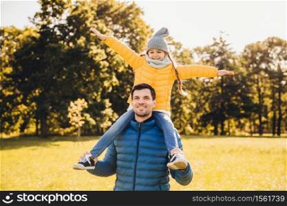 Handsome father wears anorack gives piggyback to his little funny girl, stroll together outdoor, enjoy sunshine. Attractive dad and his daughter who rides on his back, gestures, feels freedom