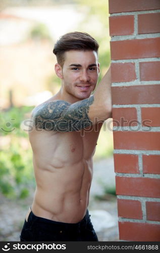 Handsome fit athletic shirtless young man with a tattoo leaning against a brick wall