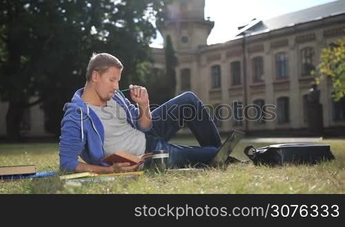 Handsome focused student sitting on the grass studying on campus lawn at college. Thoughtful male student reading book and making notes using a laptop outside the university in the park.