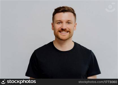Handsome good looking ginger unshaven man smiling happily at camera while standing in casual black shirt, being positive and friendly, looking with cheerful expression while posing over grey wall. Friendly positive man smiling at camera and enjoying life