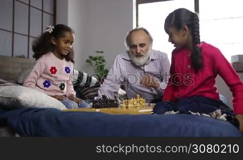 Handsome grandpa with beard teaching his cute mixed race grandaughters to play chess boards while family spending freetime together at home. Cheerful senior man helping adorable grandchildren to learn chess game indoors. Dolly shot. Slow motion.