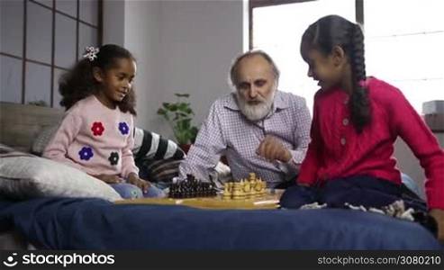 Handsome grandpa with beard teaching his cute mixed race grandaughters to play chess boards while family spending freetime together at home. Cheerful senior man helping adorable grandchildren to learn chess game indoors. Dolly shot. Slow motion.