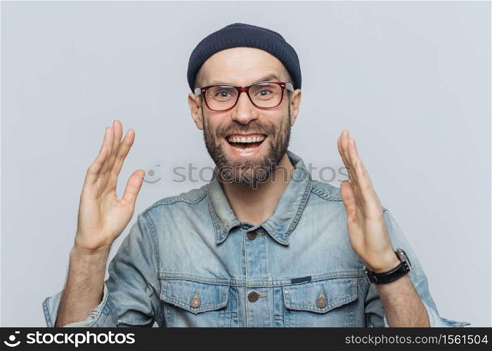 Handsome happy man raises hands with excitment, has overjoyed expression, looks with delighted face, has thick beard and mustache. Glad man in eyewear poses against white background, has fun